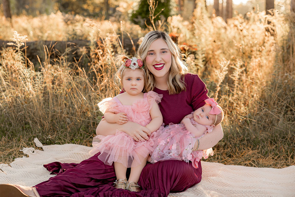 A mother sits on a picnic blanket in a park with tall grasses with her two toddler daughters in pink dresses