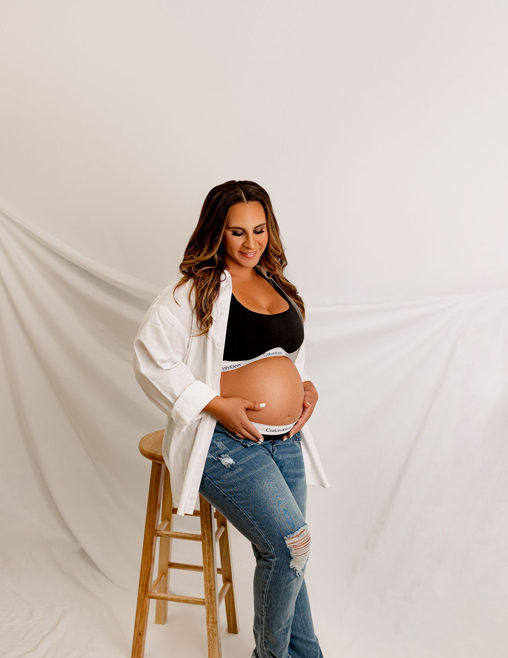 A happy mom to be stands in a studio leaning against a tall wooden stool holding and smiling down to her bump