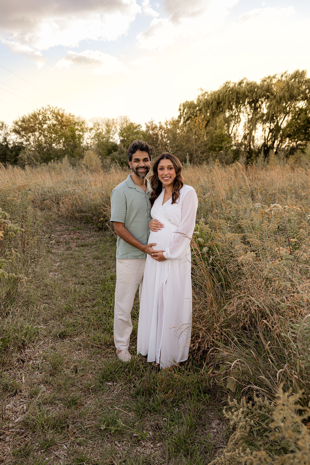 Expecting parents stand in a field of tall grasses holding the bump at sunset after meeting mothering the mother