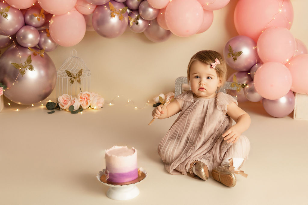 A toddler girl in a pink dress sits on a studio floor surrounded by balloons and a cake