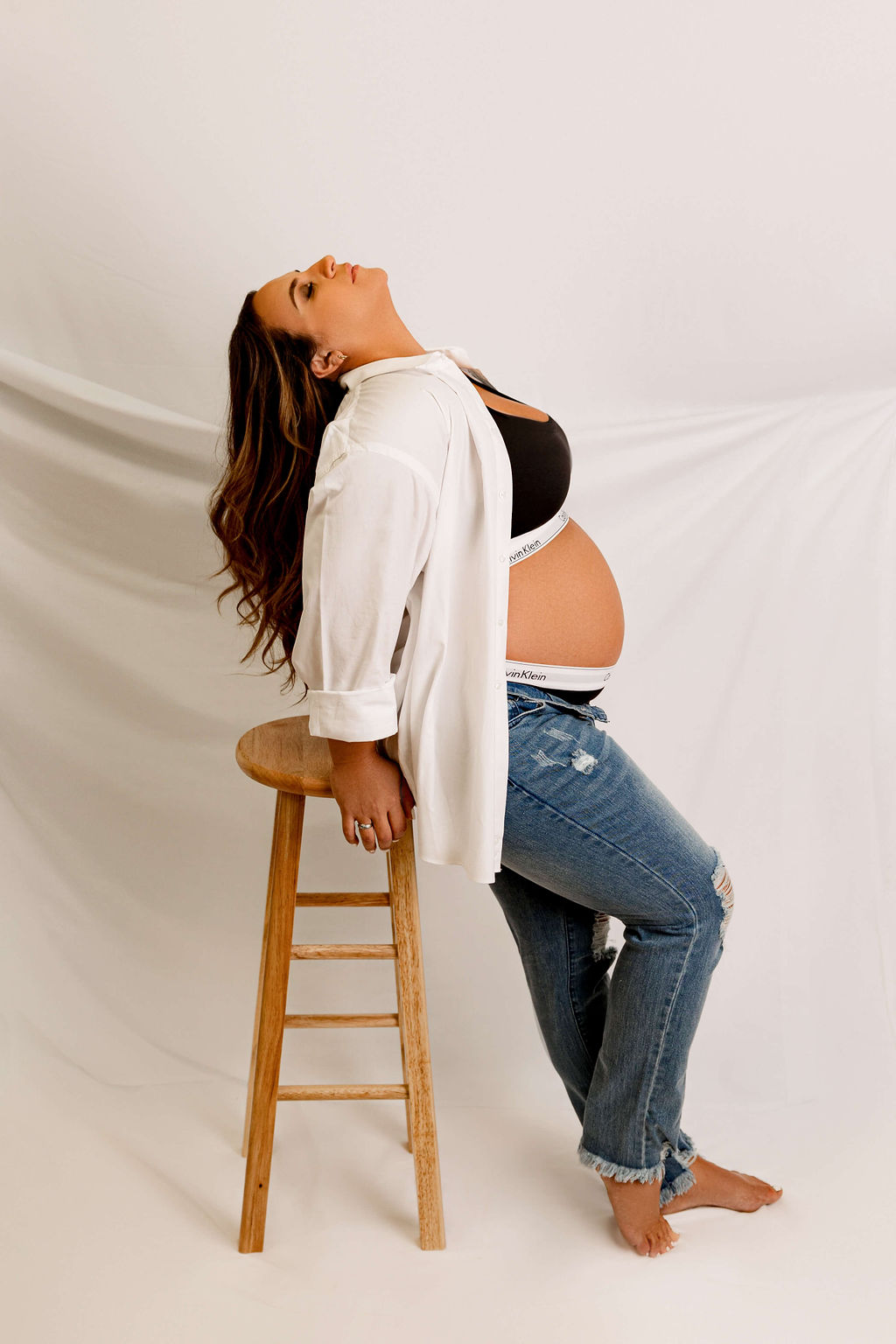 A mom to be in jeans and an unbuttoned shirt leans back on a wooden stool in a studio thanks to new life birth services