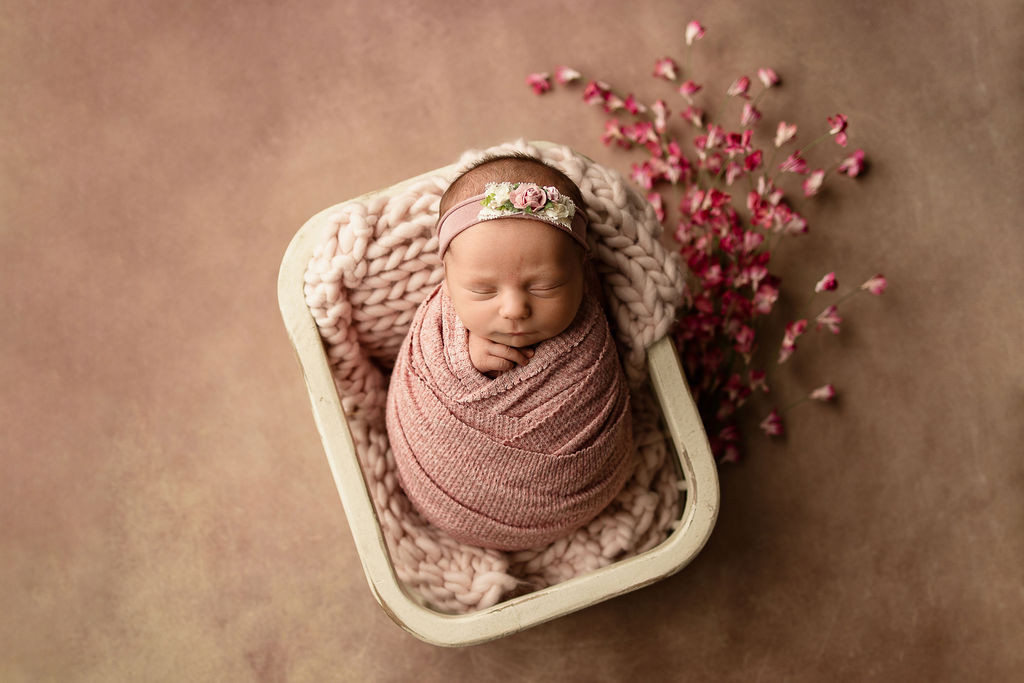 A newborn baby girl sleeps in a pink swaddle and matching headband in a wooden crate in a studio with flowers thanks to night nurse milwaukee
