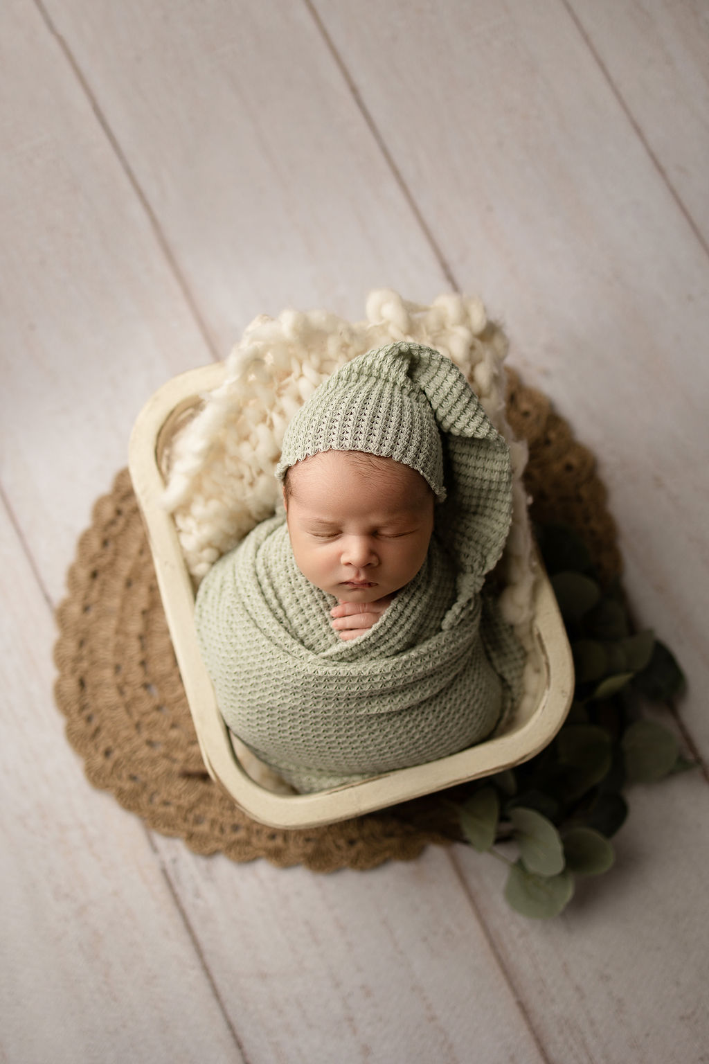 A newborn baby posed in a wooden basket sleeps in a green swaddle and night cap