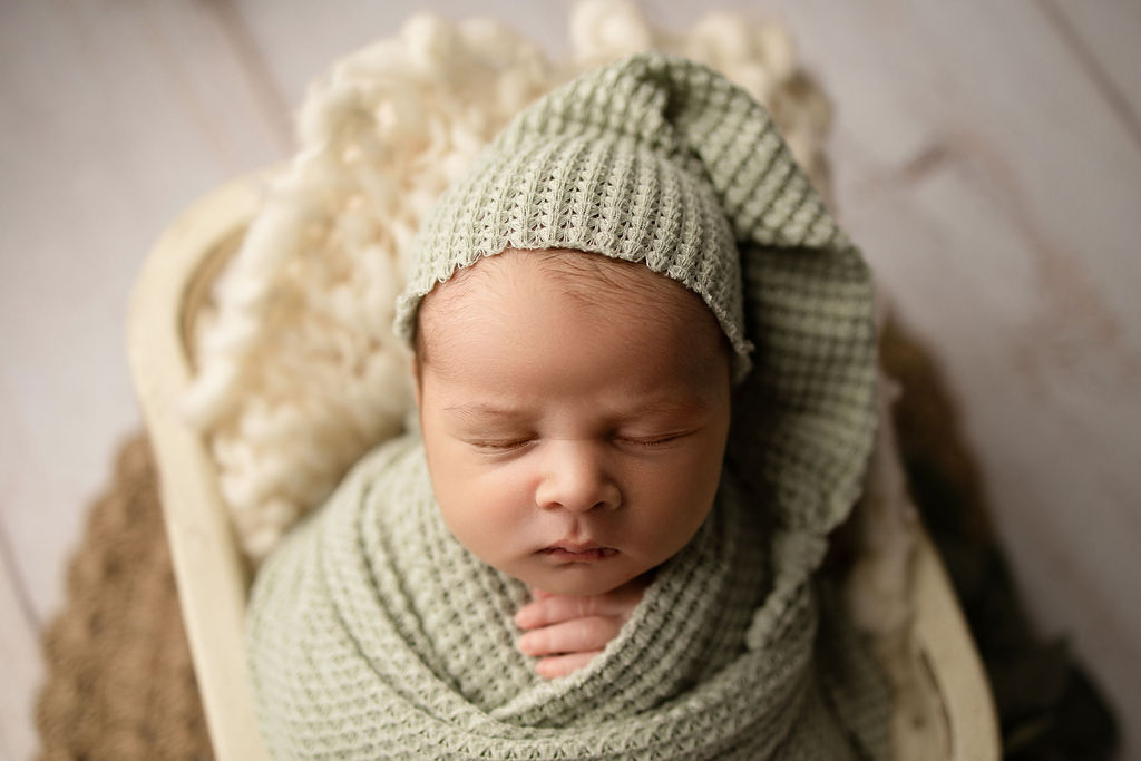 A newborn baby sleeps in a green knit nightcap and matching swaddle in a studio thanks to olive you nanny milwaukee