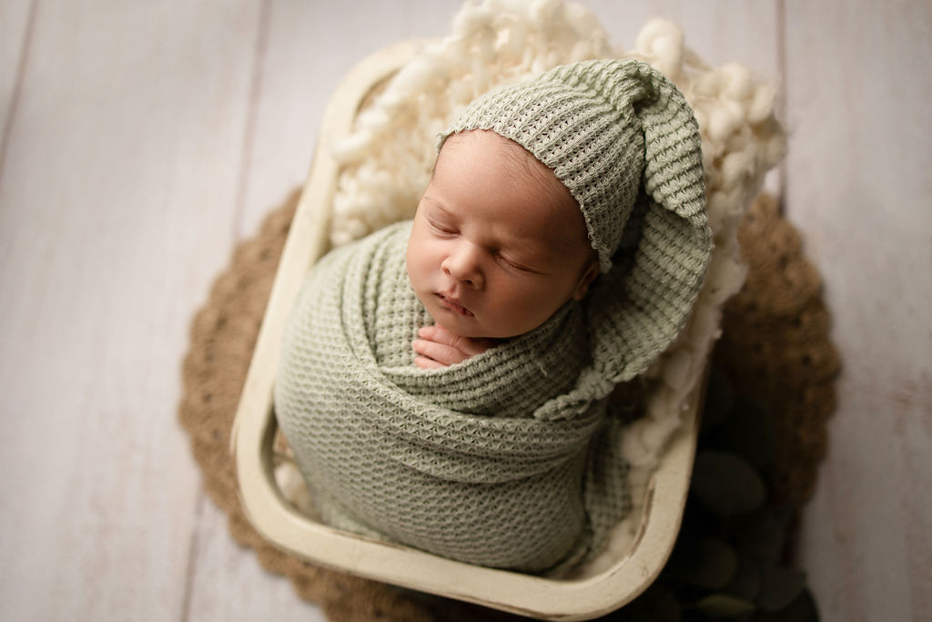 A newborn baby sleeps in a green swaddle and matching night cap in a wooden crate thanks to olive you nanny milwaukee