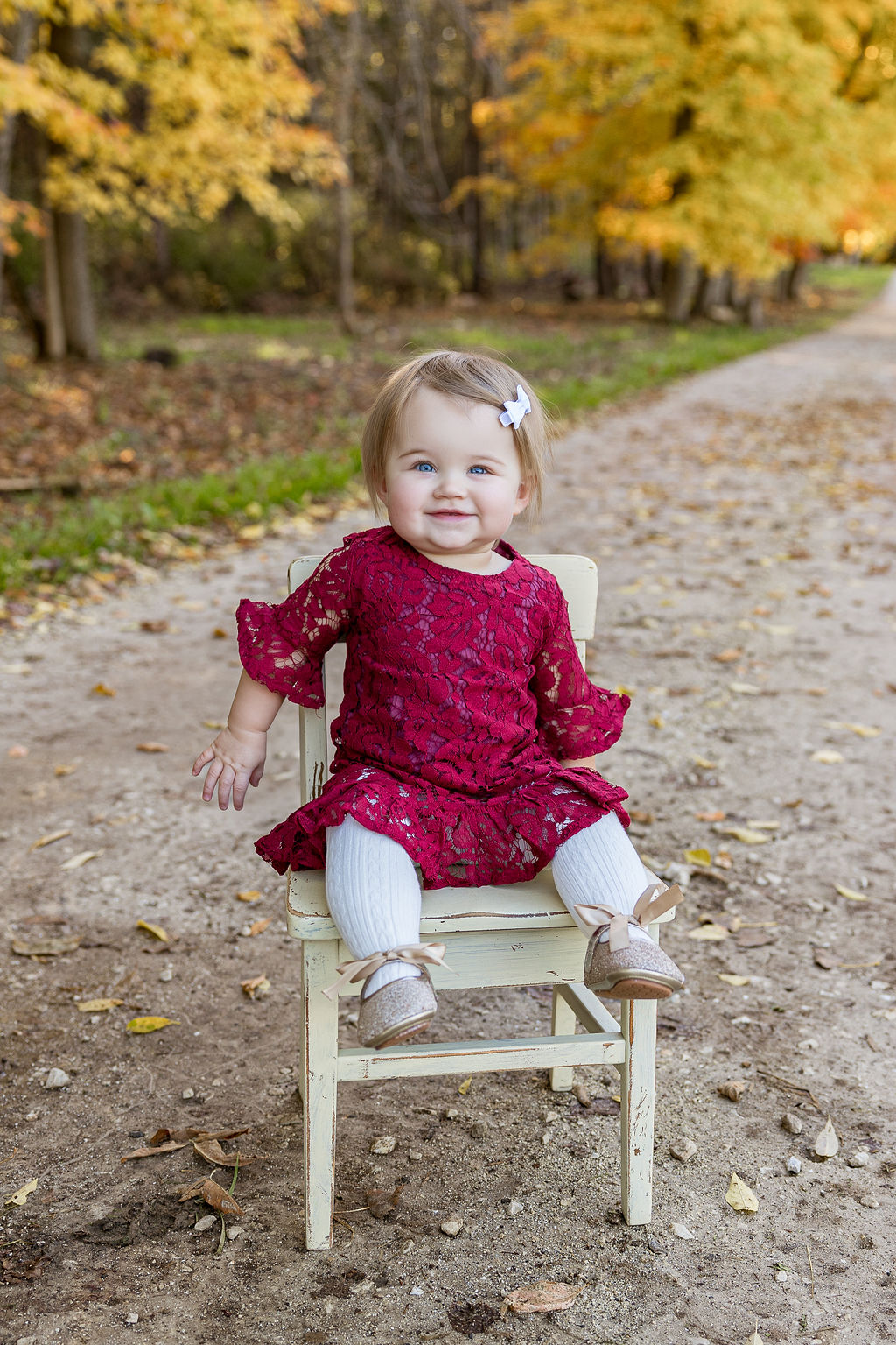 A toddler girl in a pink dress sits on a wooden chair in a park trail