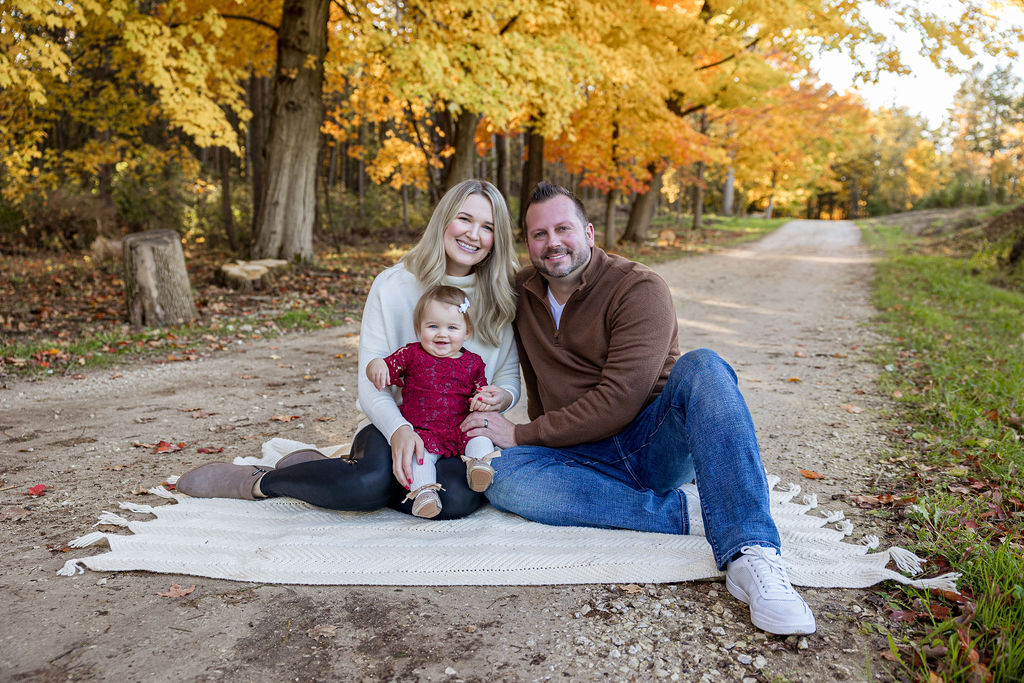 Happy parents sit on a picnic blanket with their toddler daughter in mom's lap in a park trail at fall
