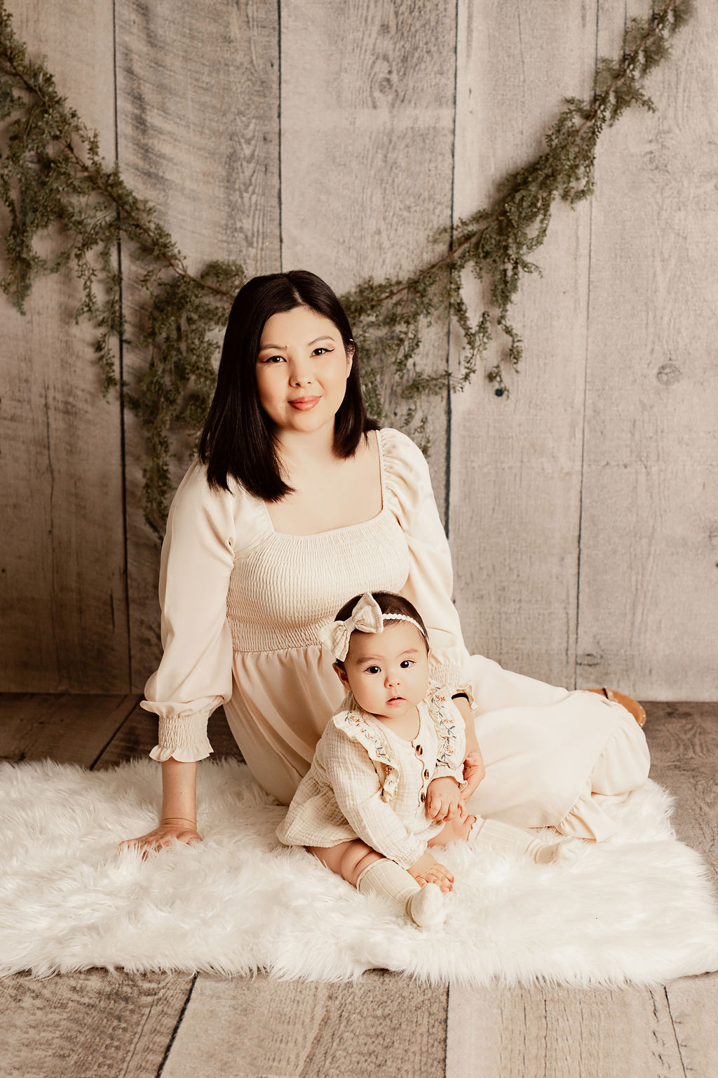 A mother smiles while sitting on the floor with her infant daughter in matching dresses on a white fur blanket thanks to pelvic floor therapy milwaukee