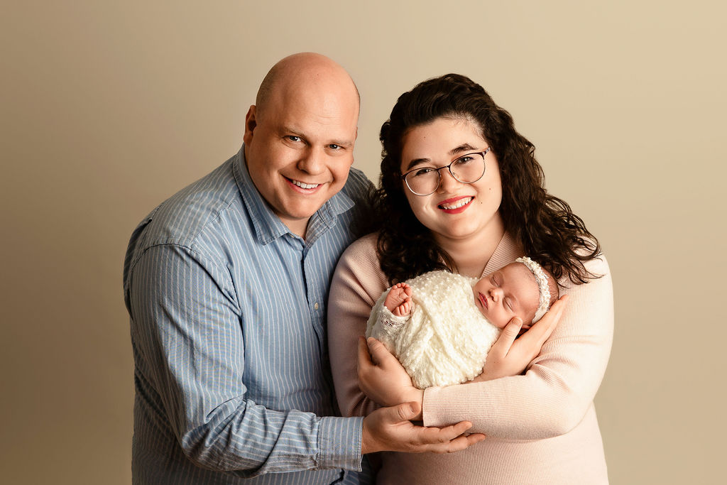 Happy parents stand in a studio cradling their sleeping newborn baby in a white swaddle