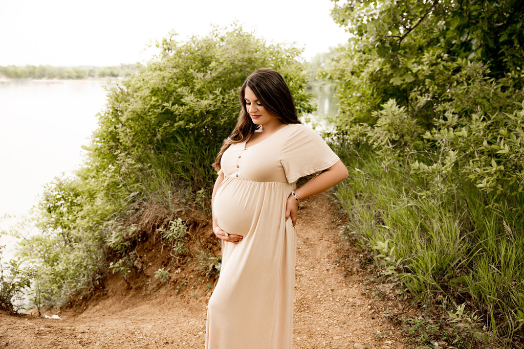 An expecting mother in a cream maternity gown stands in a lakeside trail holding her bump before a prenatal massage in milwaukee