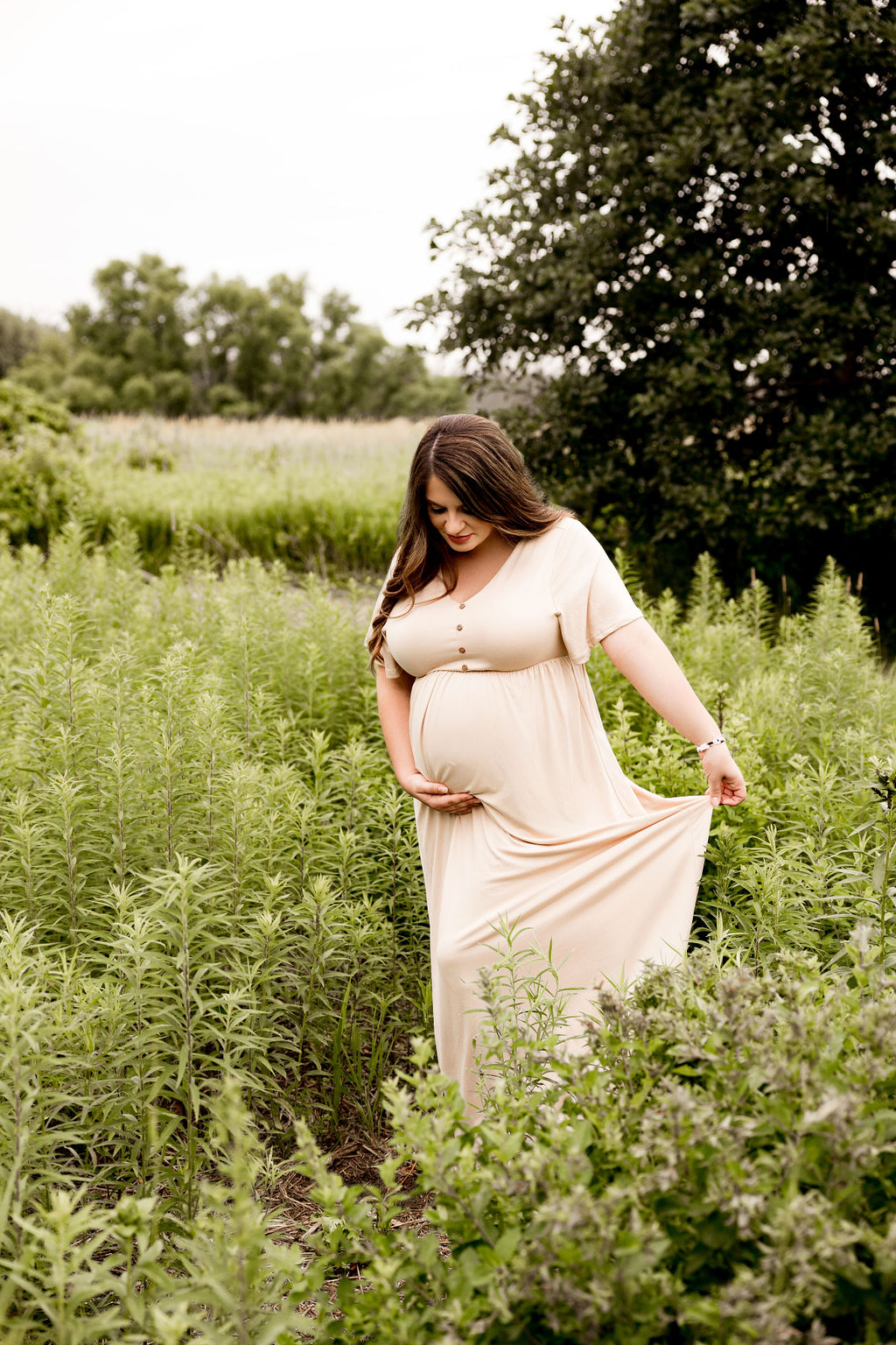 A mother to be in a cream maternity gown walks through a field of tall grasses before a prenatal massage in milwaukee