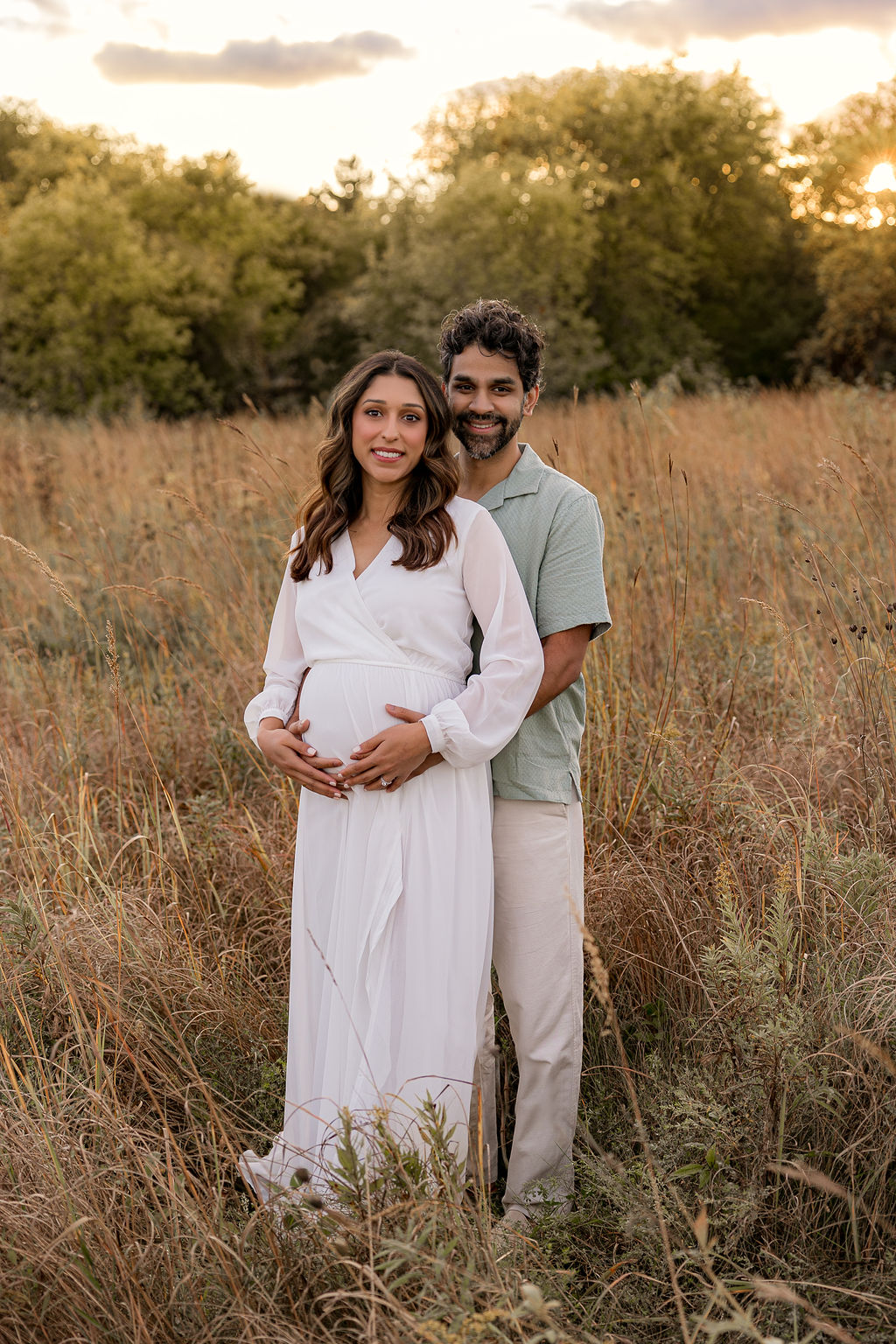 A mother and father stand in a field of tall golden grass at sunset holding the bump in a white maternity gown thanks to revitalize physical therapy wauwatosa