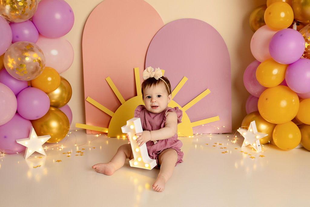 A toddler girl sits on the floor of a decorated studio playing with a light up number 1
