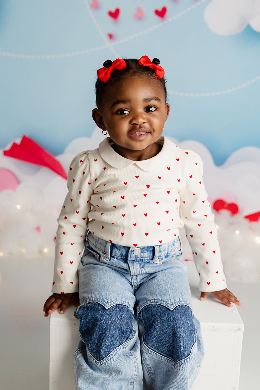 A young toddler girl in jeans and a white shirt with red hearts smiles while sitting on a wooden white box in a studio