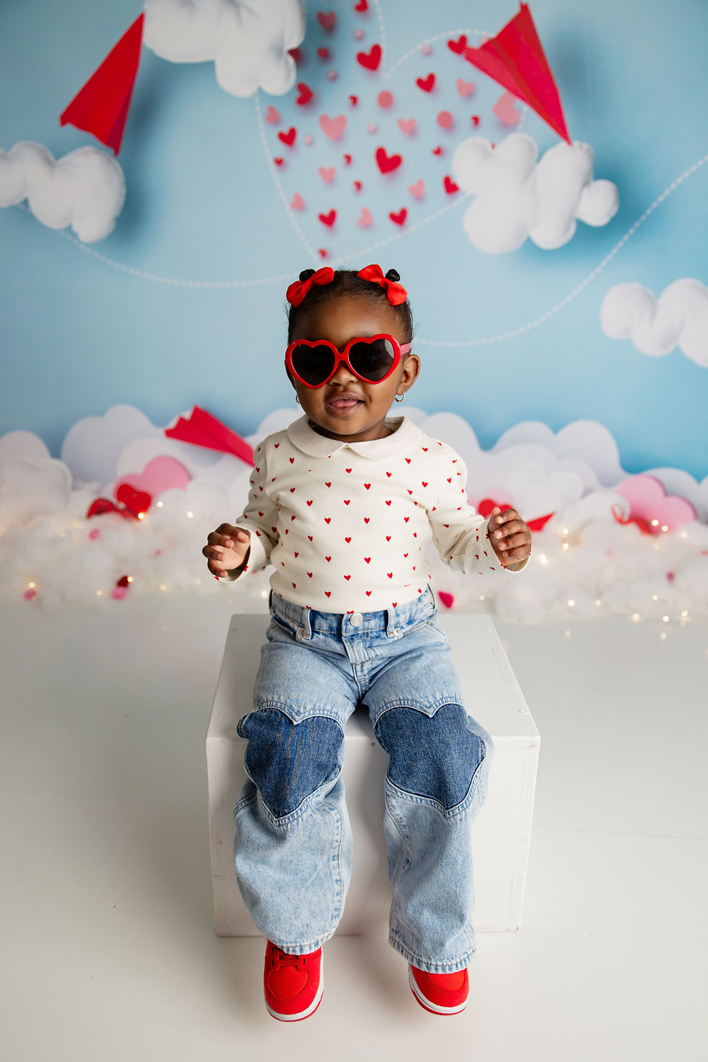 A toddler girl in red heart sunglasses sits on a white box in a studio smiling after visiting ruckus and glee