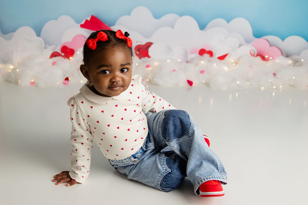 A toddler girl sits on the floor of a decorated studio in jeans and a shirt with hearts on it after visiting ruckus and glee