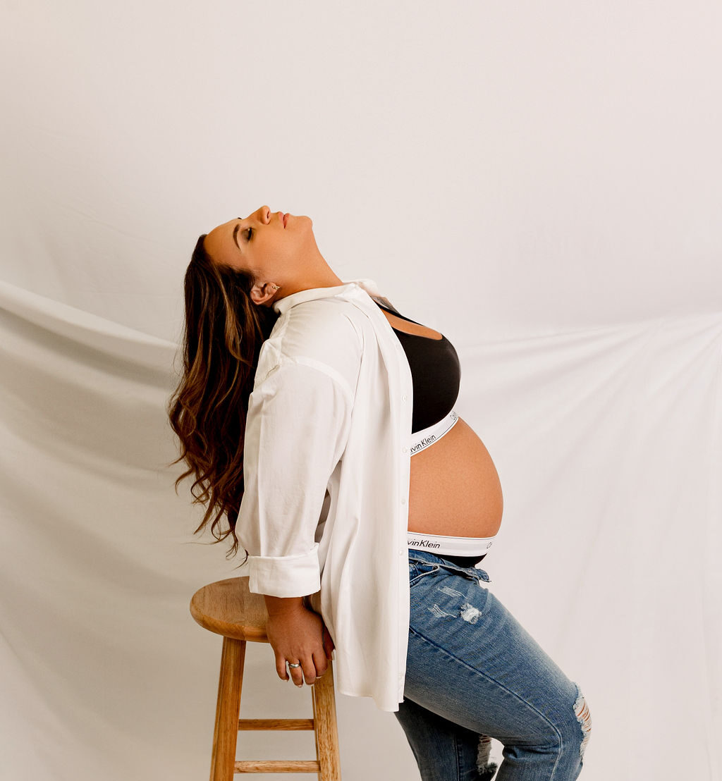 A mom to be leans against a wooden stool in a studio with her head back in jeans thanks to vios fertility milwaukee