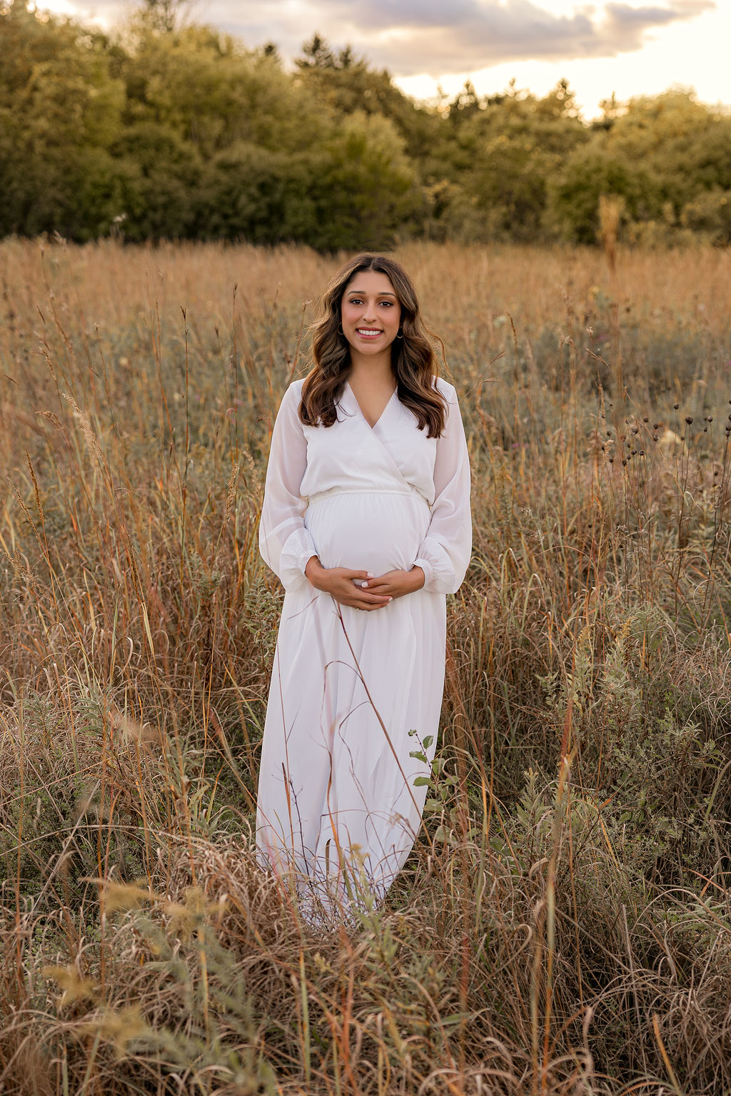 A mother to be smiles wile holding her bump and standing in a field at sunset in a white maternity gown