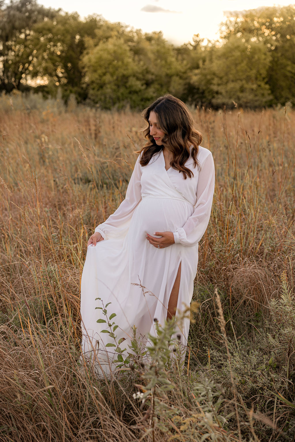 A mother to be walks through a field of tall grass while holding her dress and bump at sunset after visiting waukesha memorial hospital birthing center