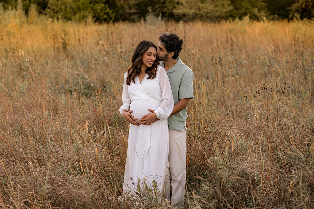 A mom to be in a white maternity gown is kissed while being hugged from behind in a field of tall golden grass at sunset after meeting waukesha memorial hospital birthing center
