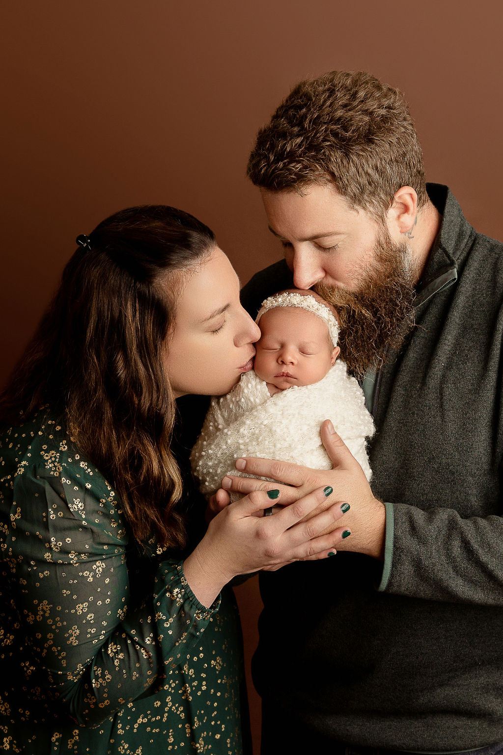 Happy parents hold and kiss their sleeping newborn baby wrapped in a white swaddle in a studio thanks to waukesha obgyn