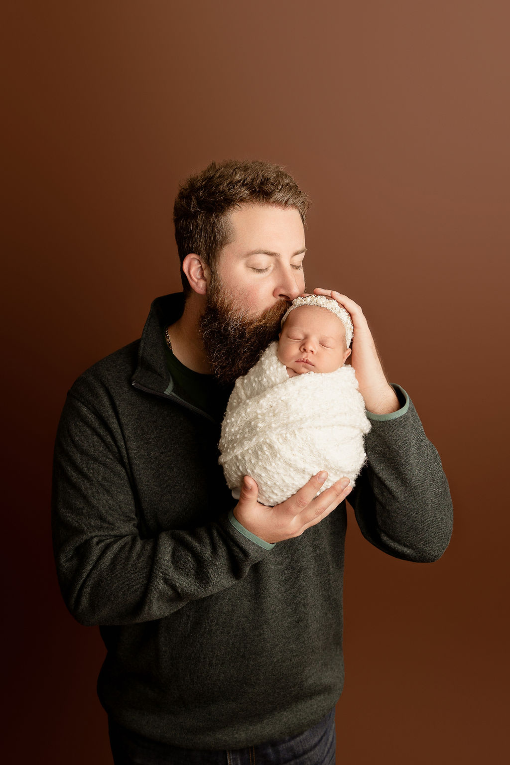 A father kisses his sleeping newborn baby while standing in a studio
