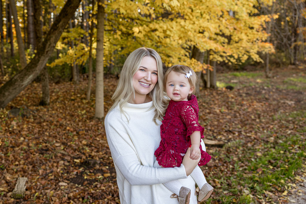 A happy mother in a white sweater stands in a park in fall while holding her toddler daughter in a red dress