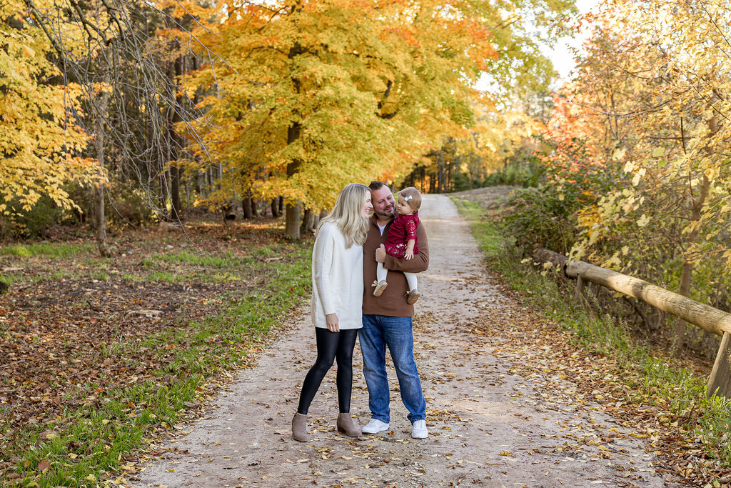 Happy parents play with their toddler daughter while walking up a park trail in fall after meeting waukesha pediatricians