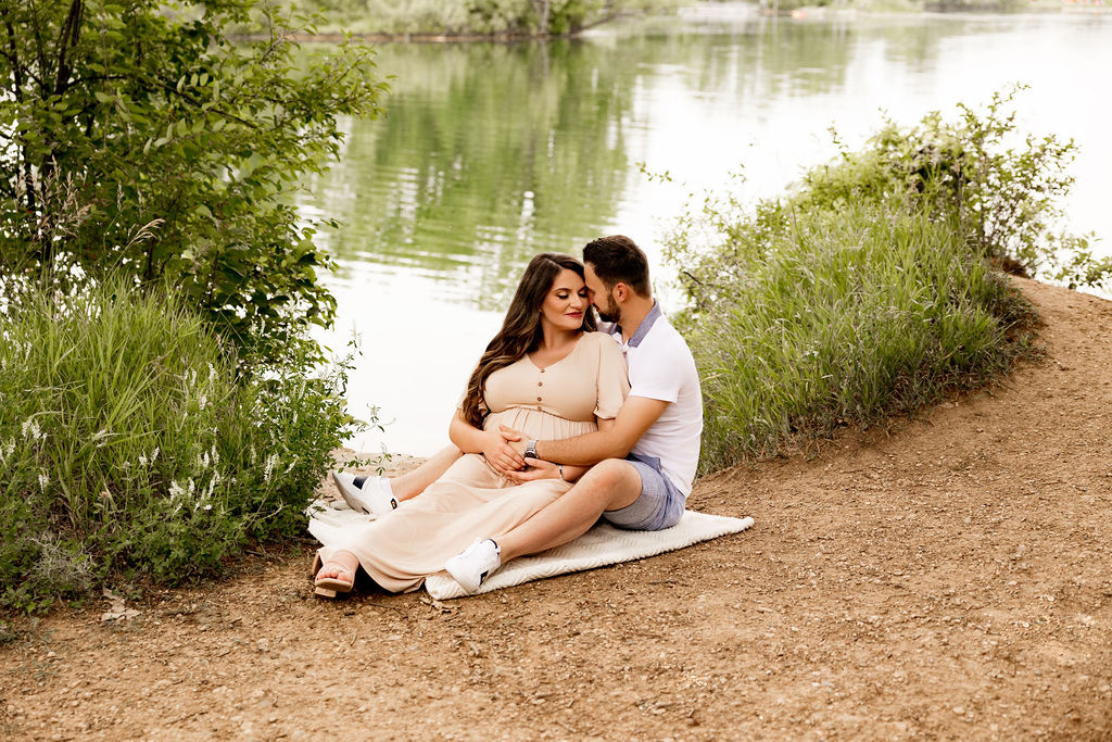 Expecting parents snuggle while sitting on a picnic blanket on the edge of a lake and holding the bump