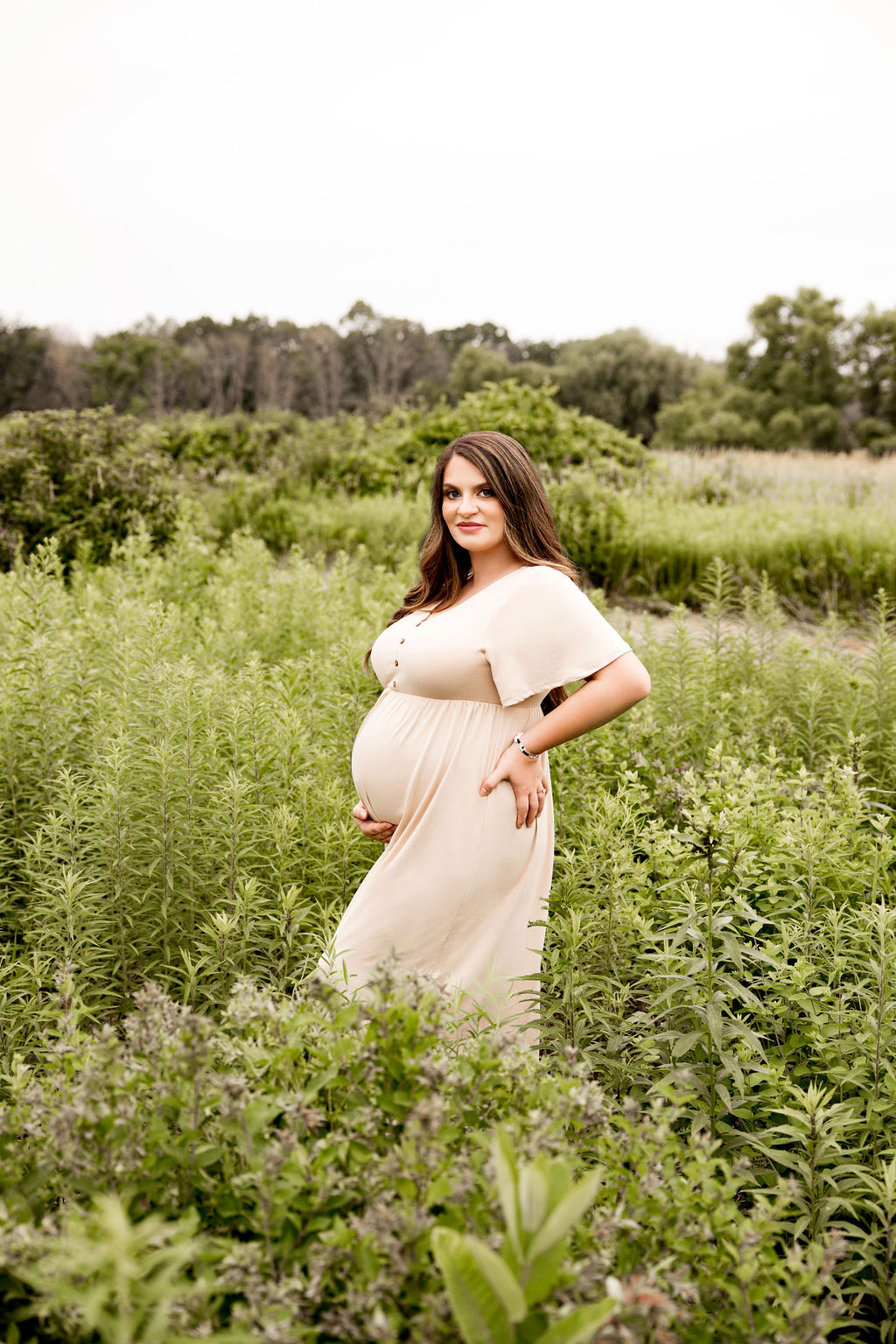 A mother to be in a cream maternity gown stands in a field of tall grasses holding the bottom of her bump after meeting with women's care center milwaukee