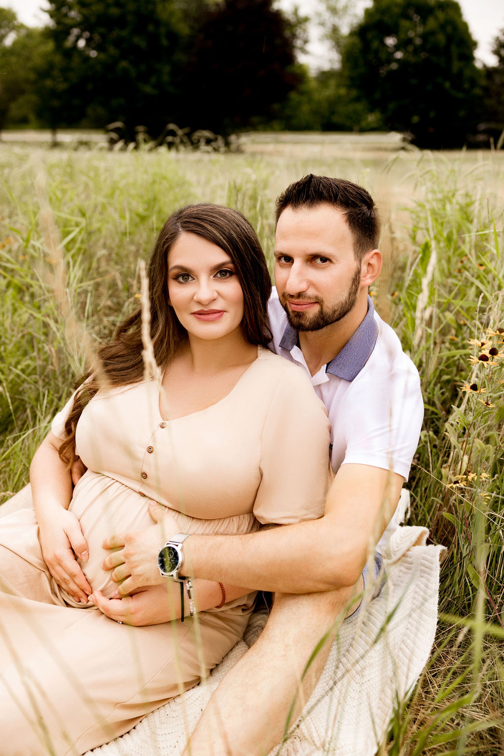 Expecting parents cuddle on a picnic blanket in a field of tall grass while holding the bump after visiting women's care center milwaukee